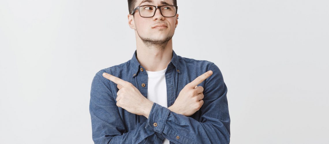 Waist-up shot of hesitant unsure attractive caucasian young man with bristle in glasses and blue shirt crossing hands on chest, pointing sideways as being doubtful what chose, looking right unsure.